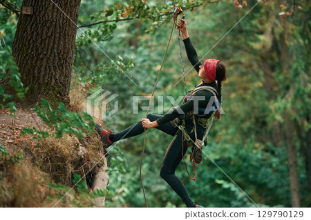 View from the side, hanging on the rope. Woman is doing climbing in the forest by the use of safety equipment View from the side, hanging on the rope. Woman is doing climbing in the forest by the use of safety equipment 129790129