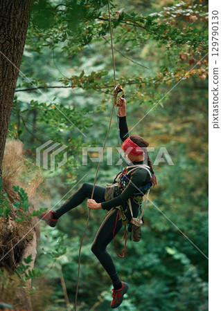 In sportive clothes, high up on the rope. Woman is doing climbing in the forest by the use of safety equipment In sportive clothes, high up on the rope. Woman is doing climbing in the forest by the use of safety equipment 129790130