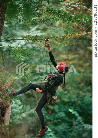 In sportive clothes, high up on the rope. Woman is doing climbing in the forest by the use of safety equipment In sportive clothes, high up on the rope. Woman is doing climbing in the forest by the use of safety equipment 129790131