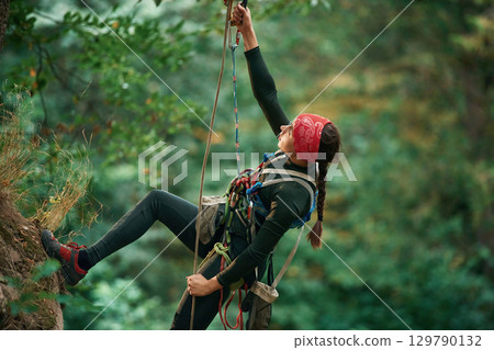 In sportive clothes, high up on the rope. Woman is doing climbing in the forest by the use of safety equipment In sportive clothes, high up on the rope. Woman is doing climbing in the forest by the use of safety equipment 129790132