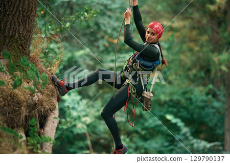 Looking to the side, hanging on the rope. Woman is doing climbing in the forest by the use of safety equipment 129790137