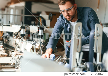 Operating the machine. Handsome man is working at the factory of creating eco boxes Operating the machine. Handsome man is working at the factory of creating eco boxes 129790670
