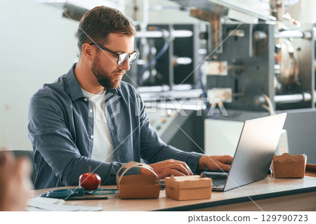 Sitting by the table. Handsome man is working at the factory of creating eco boxes 129790723