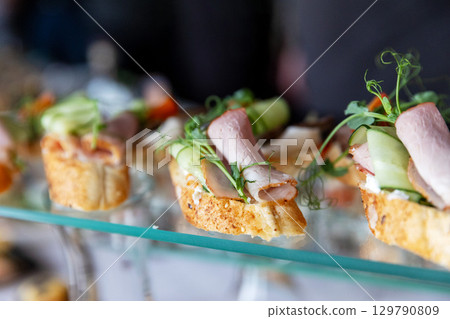 A close-up of delicious canapes on a glass tray, topped with ham, cucumber, and microgreens 129790809