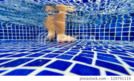 Underwater view of child feet standing in swimming pool with blue tiles 129791119