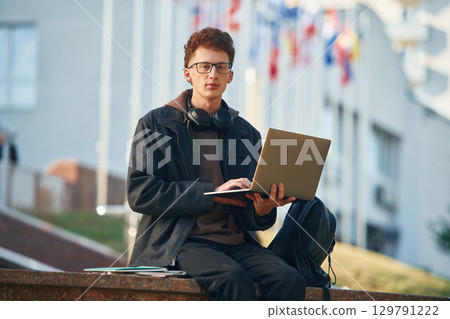 Guy is sitting and using laptop. Young student is outside the university, in black clothes Guy is sitting and using laptop. Young student is outside the university, in black clothes 129791222