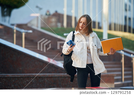Smartphone in hand. Young female student in casual clothes is outdoors Smartphone in hand. Young female student in casual clothes is outdoors 129791258
