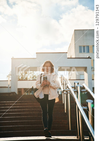 Illuminated by sunlight, holding bottle of water. Young female student in casual clothes is outdoors 129791343