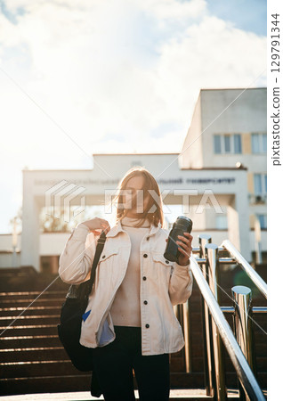 Illuminated by sunlight, holding bottle of water. Young female student in casual clothes is outdoors 129791344