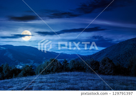 mountain landscape with forest on the hill in autumn at night. beautiful view in to the rural valley in full moon light. alpine countryside scenery under sky with clouds mountain landscape with forest on the hill in autumn at night. beautiful view in to the rural valley in full moon light. alpine countryside scenery under sky with clouds 129791397