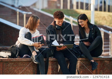 With notepad. Three young students are outside the university outdoors 129791418