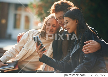 Girl is showing something in smartphone. Three young students are outside the university outdoors 129791427