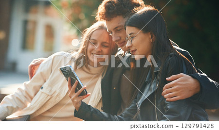 Girl is showing something in smartphone. Three young students are outside the university outdoors 129791428