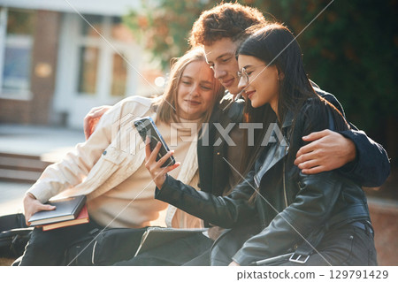 Girl is showing something in smartphone. Three young students are outside the university outdoors Girl is showing something in smartphone. Three young students are outside the university outdoors 129791429