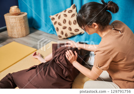 Masseuse making thai yoga massage. Woman therapist doing traditional massage treatment for female patient indoors. Client lying on mat, meditating, enjoying hand massage in calm atmosphere. Masseuse making thai yoga massage. Woman therapist doing traditional massage treatment for female patient indoors. Client lying on mat, meditating, enjoying hand massage in calm atmosphere. 129791756
