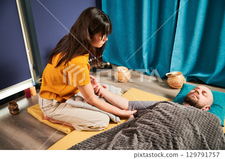 Masseuse making thai yoga massage. Woman therapist doing traditional massage treatment for male patient indoors. Bald client lying on mat, meditating, enjoying hand massage in calm atmosphere. 129791757