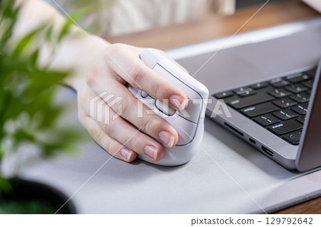 A person's hand comfortably gripping an ergonomic vertical mouse next to a laptop, promoting wrist health and efficient digital work in a modern office setup A person's hand comfortably gripping an ergonomic vertical mouse next to a laptop, promoting wrist health and efficient digital work in a modern office setup 129792642