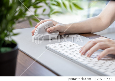 Female hand using a white vertical ergonomic mouse on a grey desk pad with a keyboard, emphasizing comfortable computing and productivity in a modern work environment Female hand using a white vertical ergonomic mouse on a grey desk pad with a keyboard, emphasizing comfortable computing and productivity in a modern work environment 129792658
