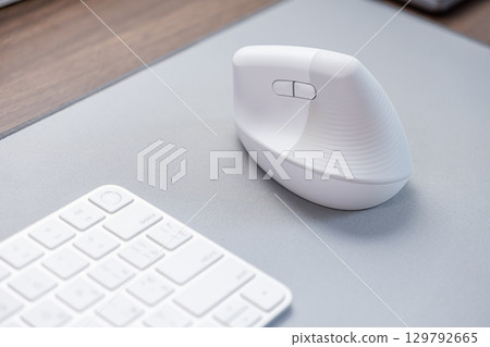 Modern white ergonomic vertical computer mouse sits on a grey desk mat next to a keyboard, emphasizing comfortable workspace setup and digital user health 129792665