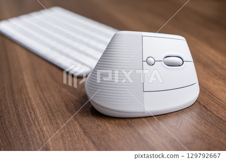 Ergonomic white vertical computer mouse on a wooden desk with a blurred keyboard, promoting wrist health, comfort, and productivity for modern professional work 129792667