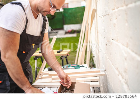 Mature male carpenter using electric screwdriver on a wooden plank outdoors 129793564