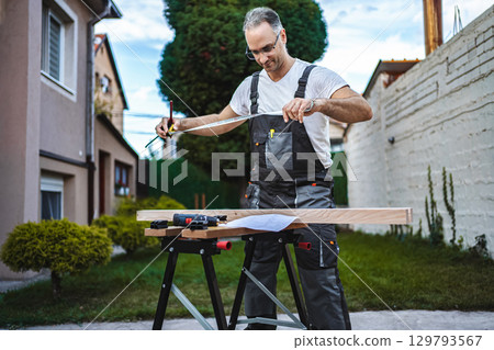 Mature male carpenter measuring wooden plank outdoors 129793567