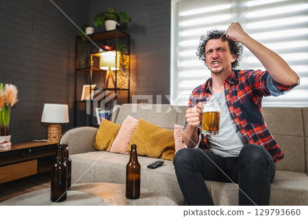 Excited sports fan celebrating with beer while watching tv at home 129793660