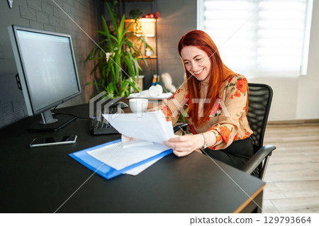 Smiling red-haired woman arranges bills on her desk Smiling red-haired woman arranges bills on her desk 129793664