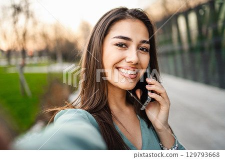 Selfie of smiling young brunette girl using mobile phone outdoors 129793668