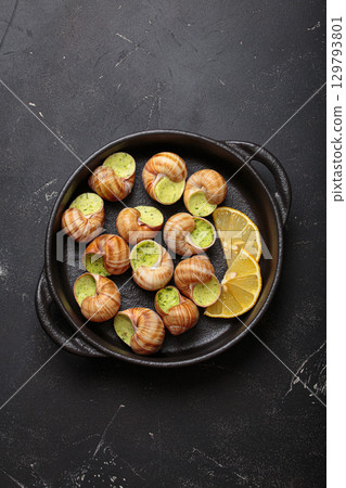 Snails escargots with garlic butter and lemon, served in a black pan on black rustic background 129793801