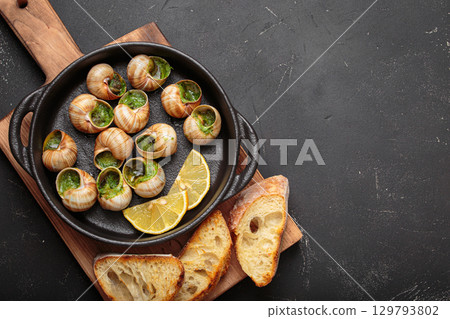 Snails escargots with garlic butter and lemon, served in a black pan with toasted bread baguette slices on black rustic background, space for text Snails escargots with garlic butter and lemon, served in a black pan with toasted bread baguette slices on black rustic background, space for text 129793802