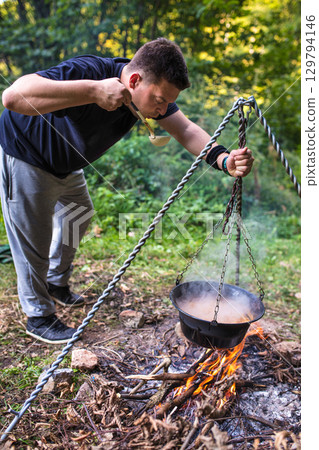 Young man tasting food from cauldron over campfire in forest 129794146