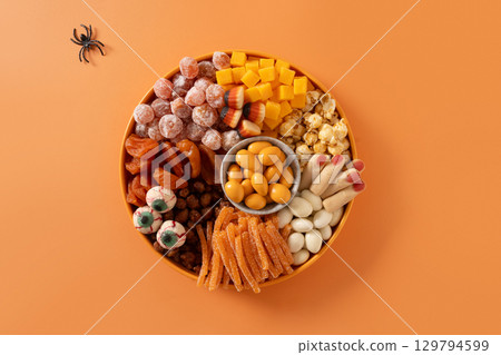 Halloween sweet charcuterie board with pumpkin, popcorn, dried fruits, color chocolate candy, spider, crackers on orange background. Festive food for kids party. Top view. Halloween sweet charcuterie board with pumpkin, popcorn, dried fruits, color chocolate candy, spider, crackers on orange background. Festive food for kids party. Top view. 129794599