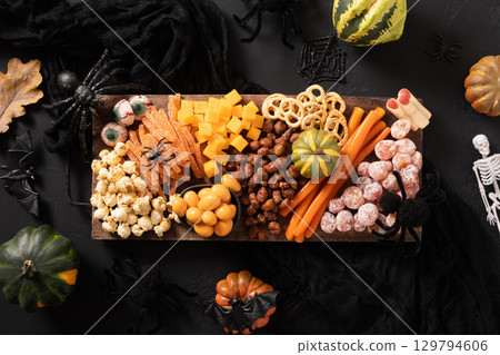 Halloween sweet charcuterie board with pumpkin, popcorn, color chocolate candy, spider, crackers with scary decoration on dark background. Festive food for kids party. View from above. 129794606