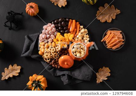 Halloween sweet charcuterie board with pumpkin, popcorn, orange chocolate candy, spider, crackers on black background. Scary decoration. Festive food for kids party. View from above. Halloween sweet charcuterie board with pumpkin, popcorn, orange chocolate candy, spider, crackers on black background. Scary decoration. Festive food for kids party. View from above. 129794608