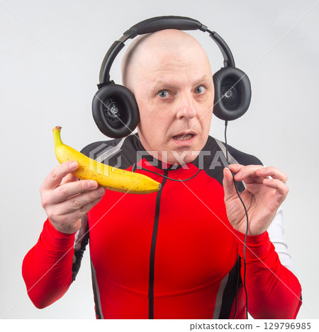 Man in red sports outfit holds banana while wearing oversized headphones in studio 129796985