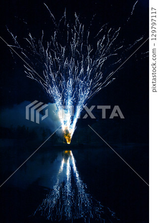 Bright fireworks illuminate the night sky over a calm lake during a festive celebration 129797117