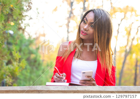 African woman writing in a notebook while enjoying a sunny outdoor setting in autumn African woman writing in a notebook while enjoying a sunny outdoor setting in autumn 129797157