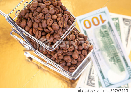 Coffee beans stacked in a shopping cart next to hundred dollar bills on a golden surface 129797315