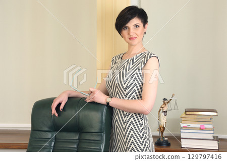 Professional woman stands confidently in an office setting near a desk filled with books 129797416