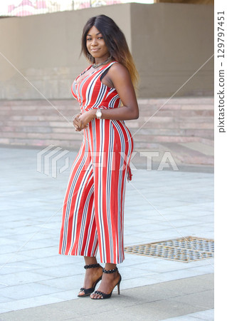 Confident African woman in stylish striped jumpsuit poses outdoors during sunny day Confident African woman in stylish striped jumpsuit poses outdoors during sunny day 129797451