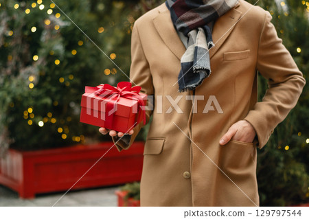 Mens wearing beige coat holding Christmas red gift box near Xmas tree at city market. Xmas shopping and happy holiday. Outdoors. Close up. Mens wearing beige coat holding Christmas red gift box near Xmas tree at city market. Xmas shopping and happy holiday. Outdoors. Close up. 129797544