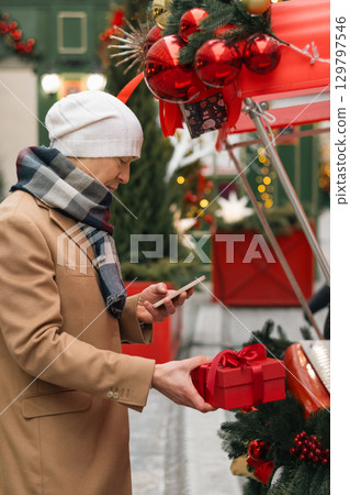 Mans buying red gift using smartphone at Christmas fairy. Xmas shopping. Side view. Holiday spirit. Close up. Mans buying red gift using smartphone at Christmas fairy. Xmas shopping. Side view. Holiday spirit. Close up. 129797546