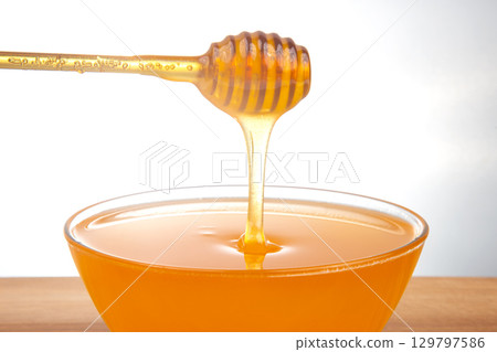 Smooth honey dripping from a wooden dipper into a clear bowl, perfect for sweetening beverages Smooth honey dripping from a wooden dipper into a clear bowl, perfect for sweetening beverages 129797586