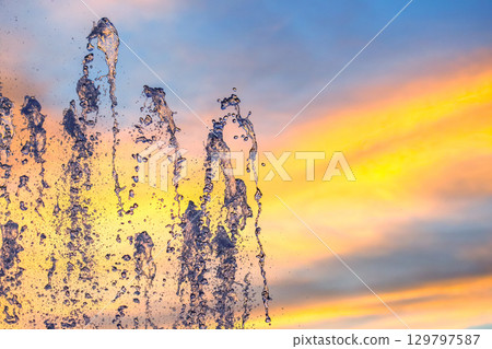 Water fountain splashes against a vibrant sunset sky with colorful clouds Water fountain splashes against a vibrant sunset sky with colorful clouds 129797587