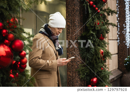 Adult handsome man with mobile phone in his hand and gifts leaves luxury store. Xmas shopping. Man Checking Phone by festive Christmas decorations. Side view. Adult handsome man with mobile phone in his hand and gifts leaves luxury store. Xmas shopping. Man Checking Phone by festive Christmas decorations. Side view. 129797928