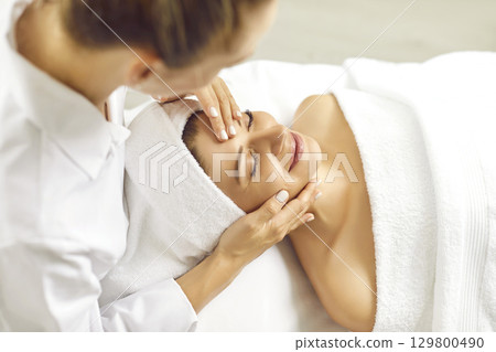 Close up photo of pretty young woman lying with closed eyes and having face or head massage in spa 129800490