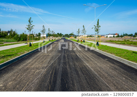 Newly paved asphalt road with sidewalks, young trees, and street lighting in modern urban development 129800857