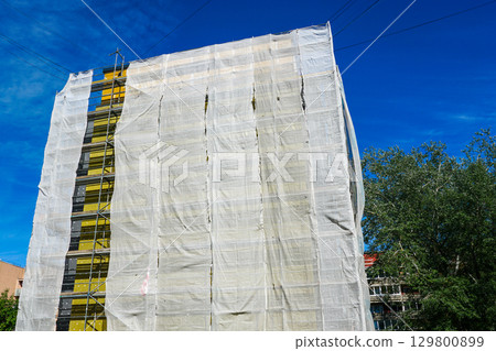 Facade thermal insulation of apartment building with scaffolding and protective mesh covering Facade thermal insulation of apartment building with scaffolding and protective mesh covering 129800899