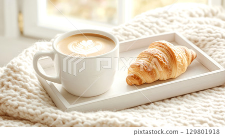 Breakfast tray with latte art coffee and fresh croissant by a sunlit window 129801918